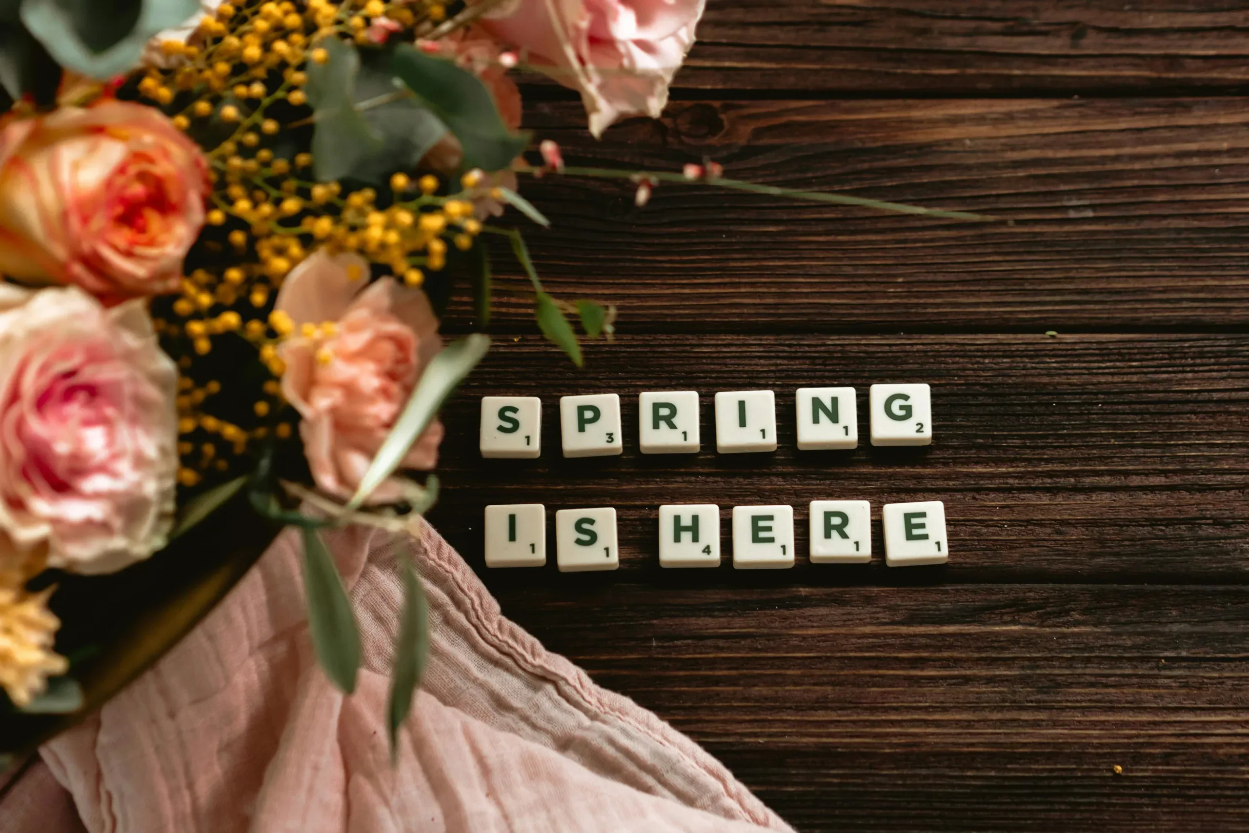 scrabble pieces spell out "spring is here" aside flowers